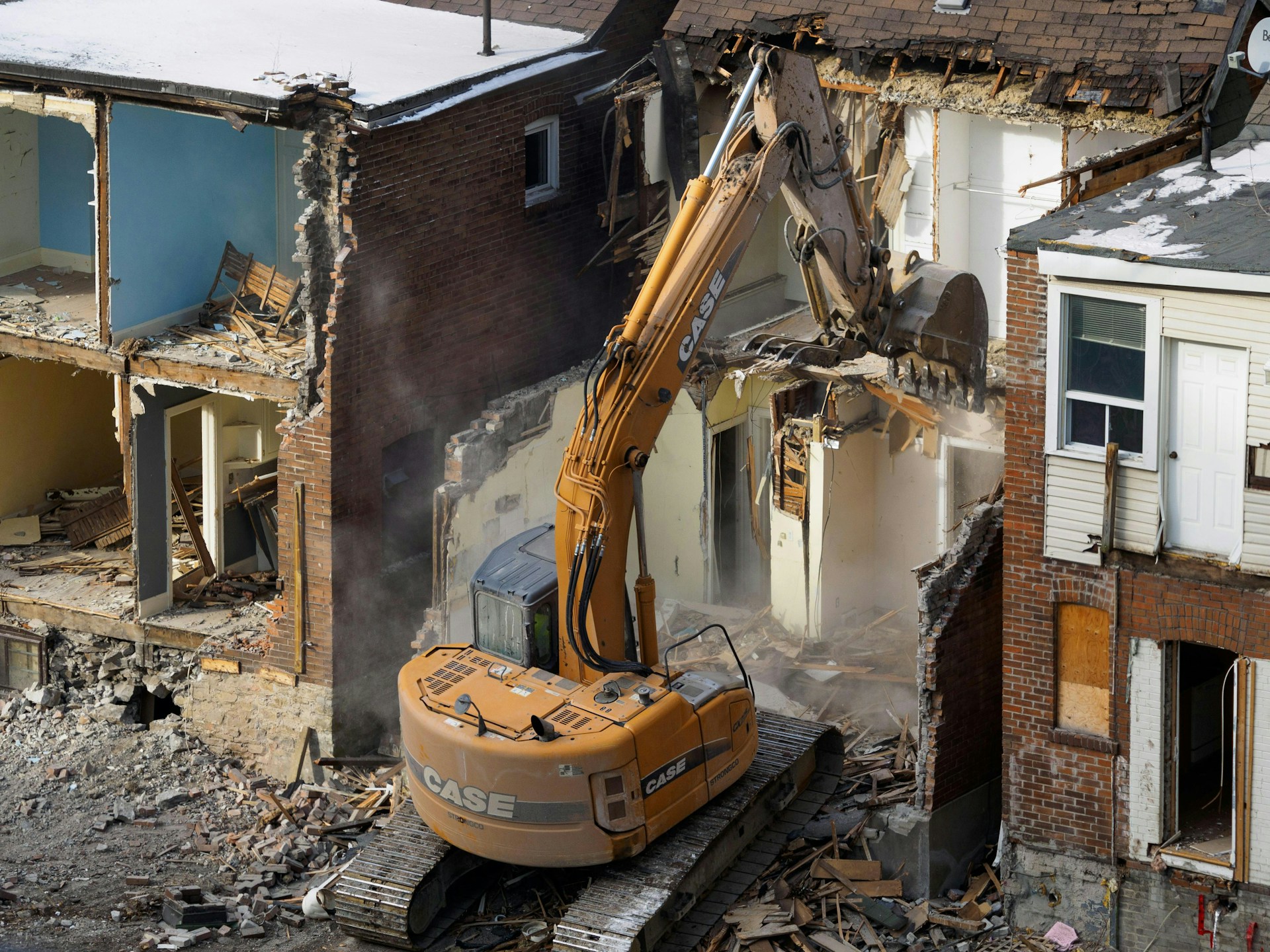 Yellow CASE excavator demolishing a brick building, exposing interior walls and rubble around it.