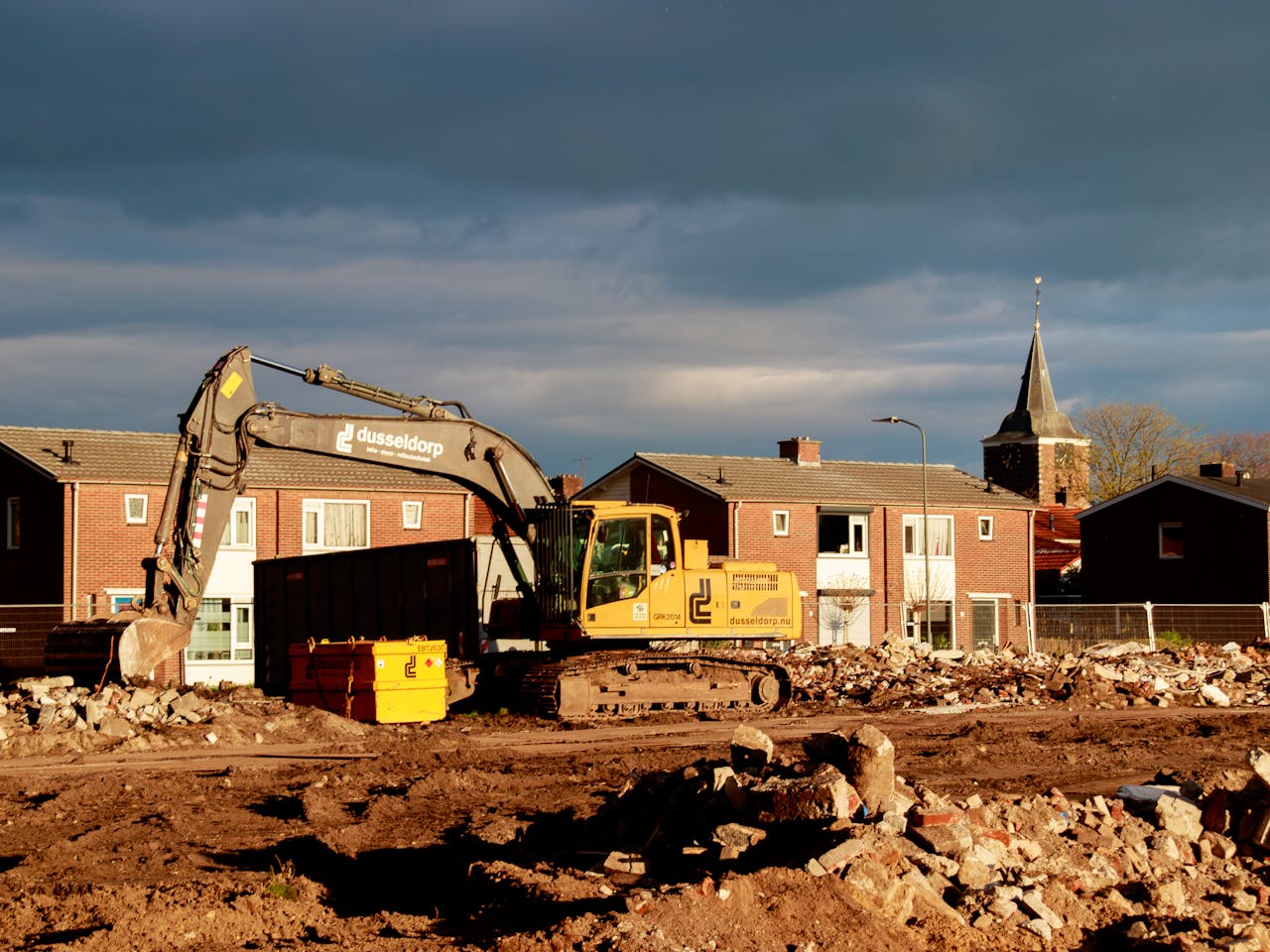 Construction site with a yellow excavator moving rubble in a residential area, brick houses and a church steeple in the background