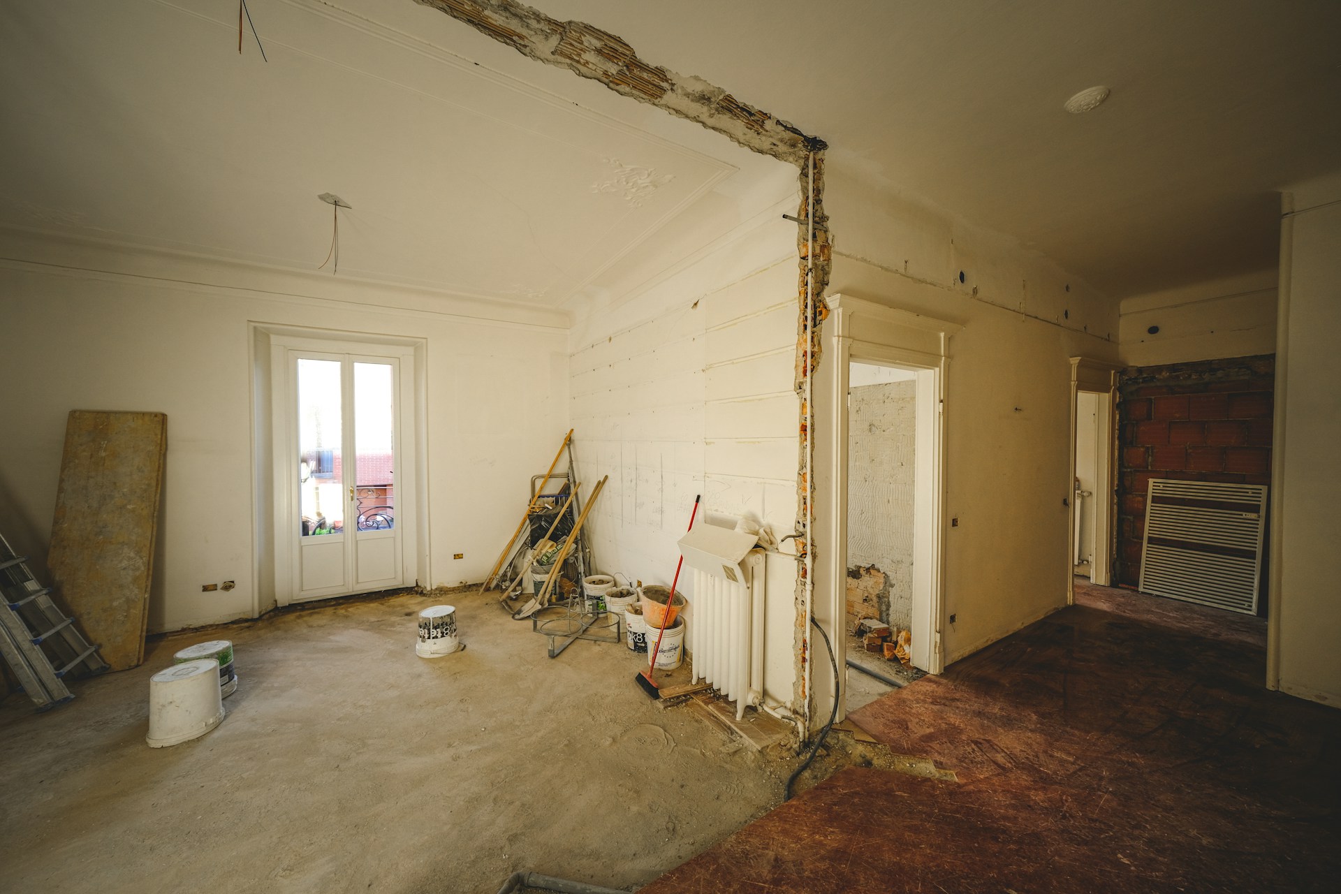 Interior under renovation with an exposed brick column, peeling plaster, and scattered buckets and tools on the floor.