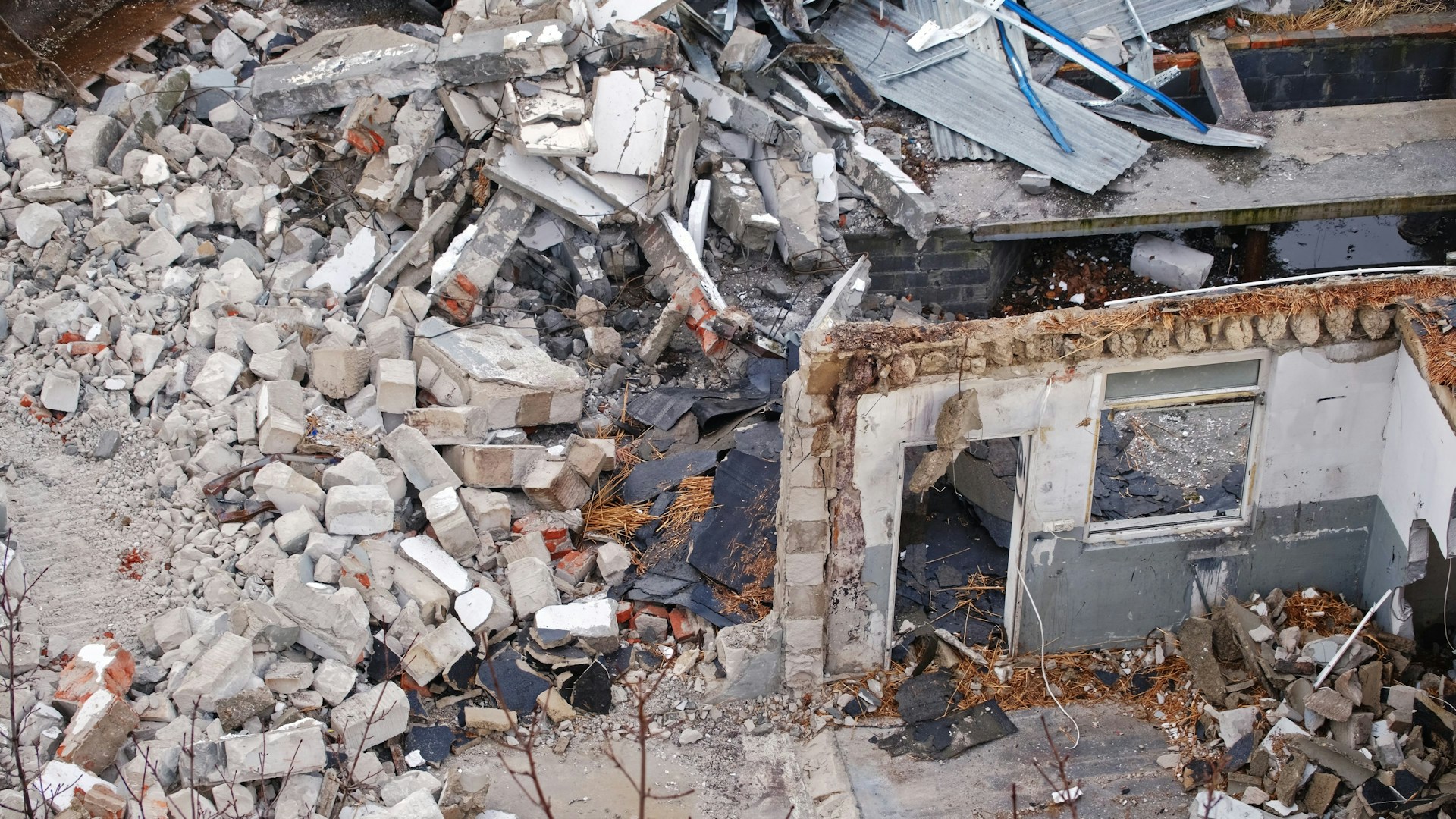 Piles of concrete rubble and bricks surround a collapsed building, exposing broken walls and window openings in the ruins.